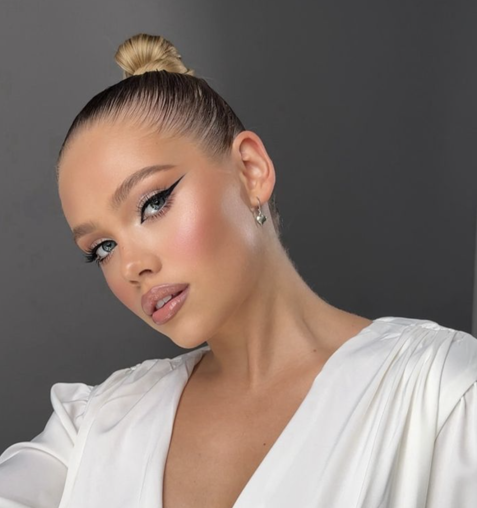 The portrait showcases a woman with a sleek, high bun hairstyle and a polished makeup look. She has a winged eyeliner, icy shimmer eyeshadow, and long, lush lashes. Her complexion glows with a soft highlighter, and her lips are tinted with a subtle pink gloss. She exudes a modern, chic vibe, accented by minimalist jewelry.
